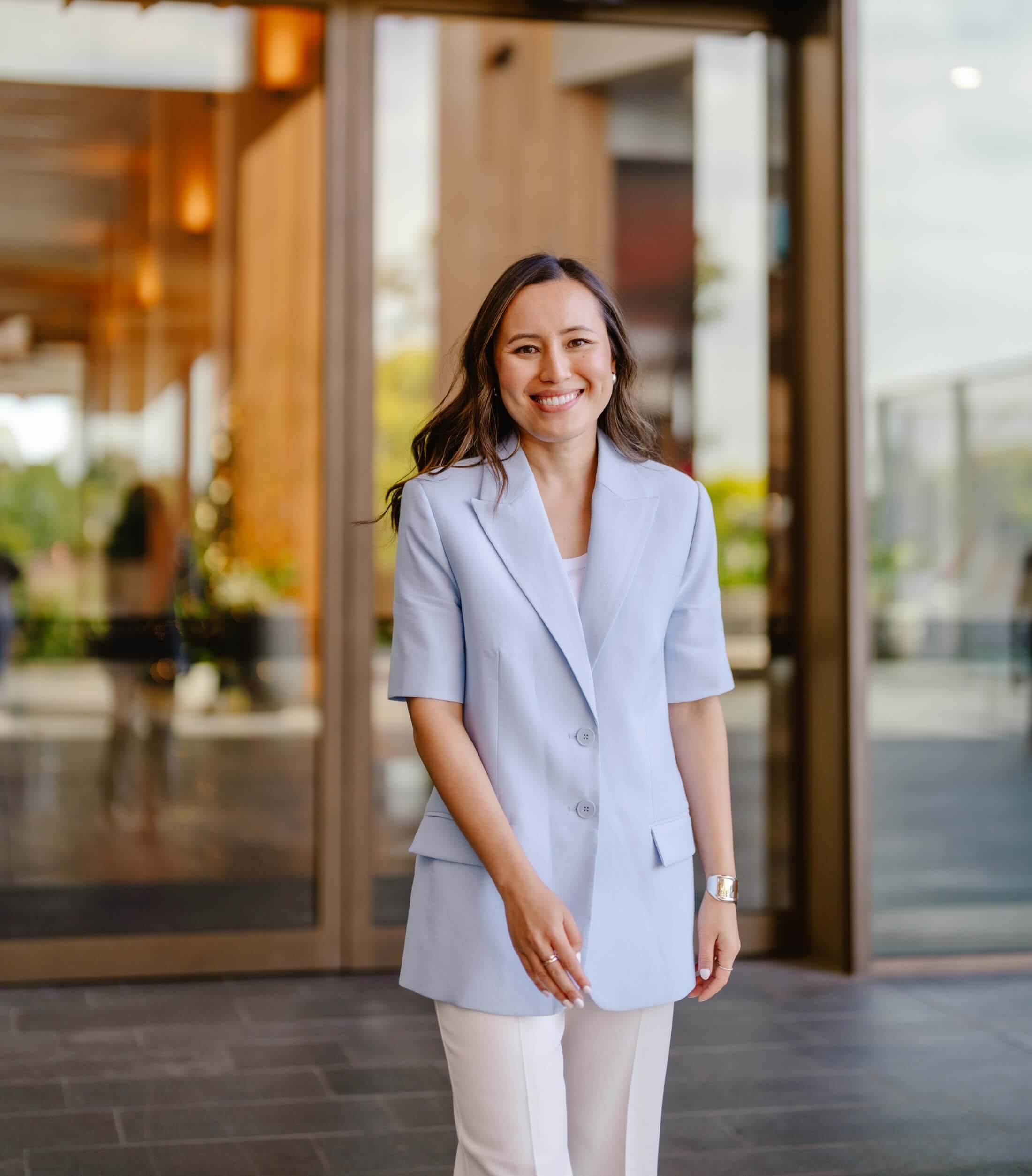 Gulinka, Melbourne personal stylist, smiling in a powder-blue blazer and white trousers outside a modern building